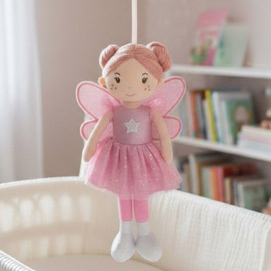 A pink plush fairy baby mobile or hanging doll with a glittery pink dress, translucent wings, and pink hair buns, suspended in front of a softly blurred nursery background with a crib and bookshelf.