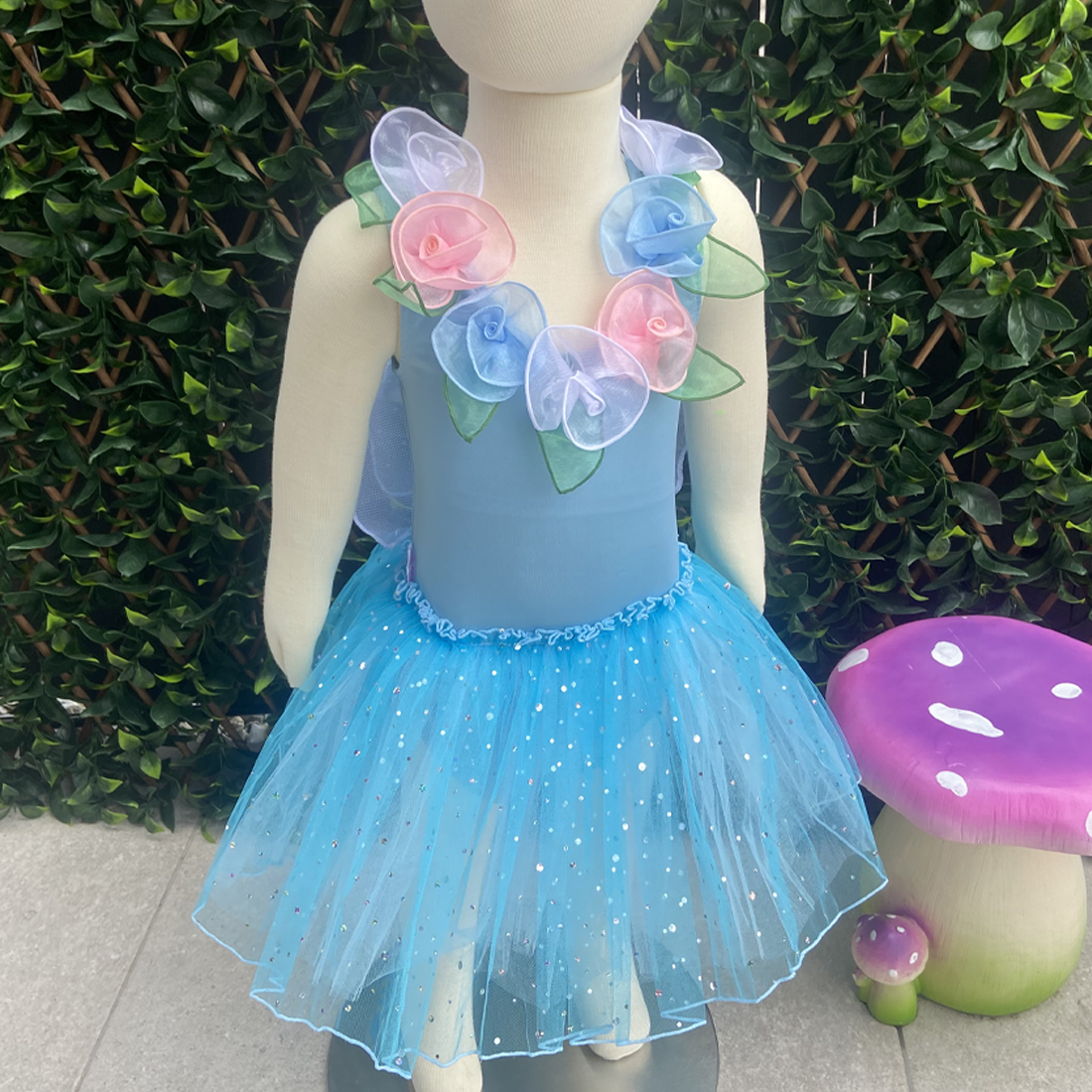 A toddler-sized blue fairy dress with a glittering skirt, handmade flowers, and decorative wings displayed on a mannequin. The mannequin is posed with a green mushroom and a pink fairy tale mushroom in the background.