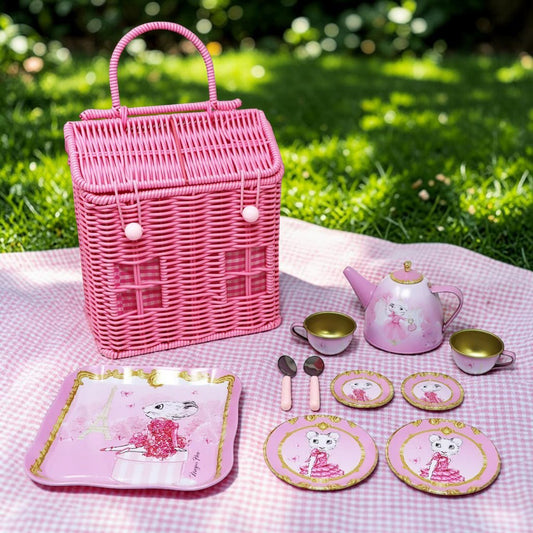 Pink wicker picnic basket set with matching tea set, plates, spoons, and cups arranged on a pink gingham blanket in a grassy outdoor setting.