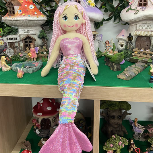A plush mermaid doll with pink hair and a gold and pink sequined body, displayed among other toy figurines.