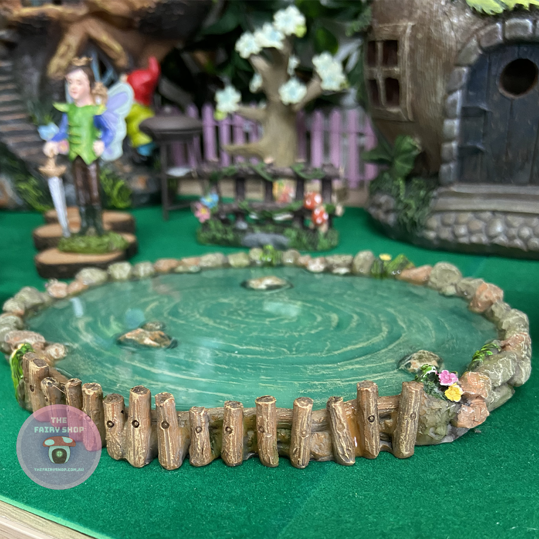 A round, hand-painted polyresin fairy garden pond with a green and natural-colored border and a central pond filled with water. Decorative elements include a fairy figure, a birdhouse, and flowers.
