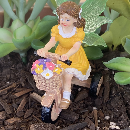 Fairy figurine in a yellow dress riding a scooter with a basket of flowers in a garden setting.