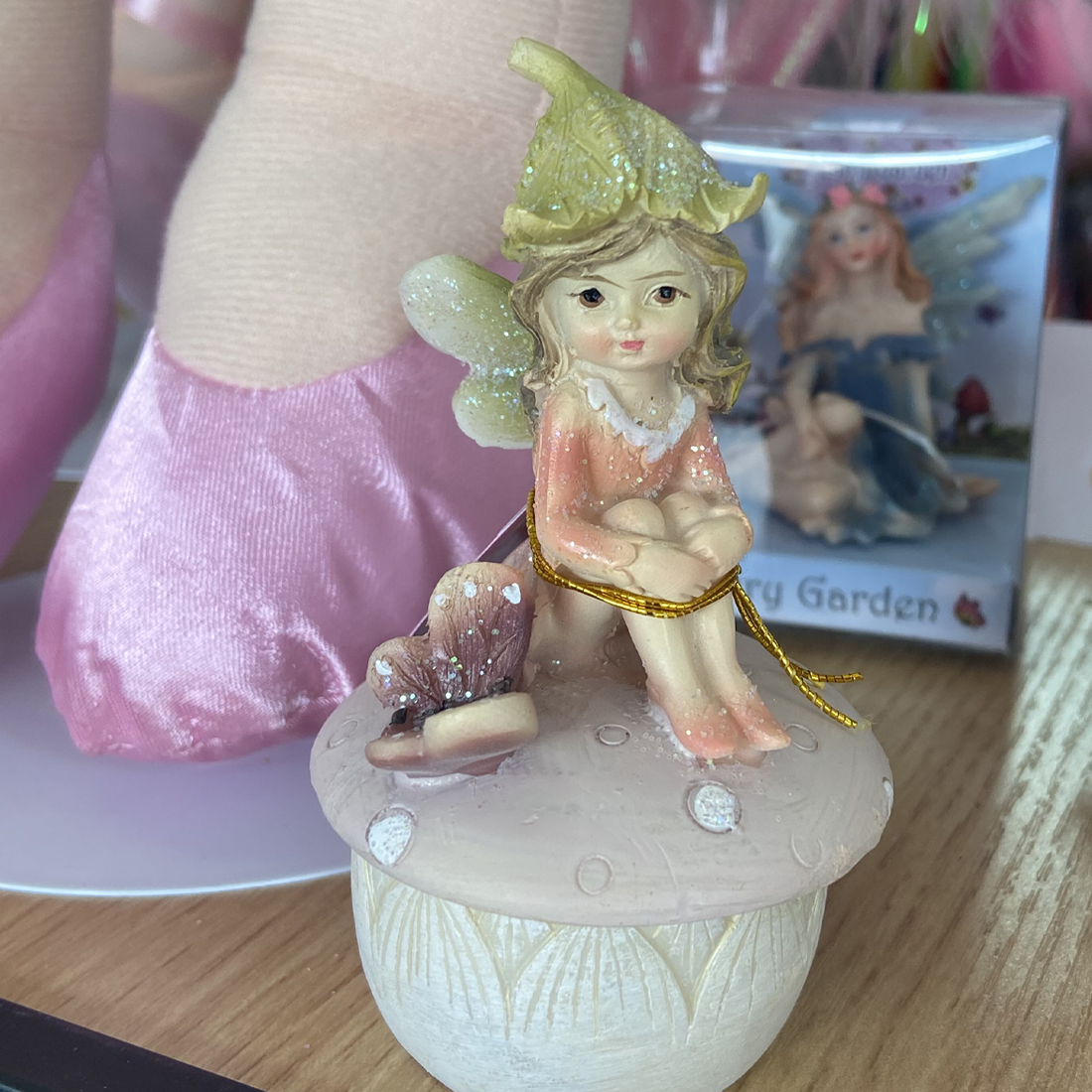 A polyresin trinket box featuring a hand-painted fairy sitting on top of a box in the shape of a mushroom with a pink lid with floral patterns.