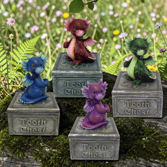 Four small fantasy dragon Tooth Chest figurines displayed outdoors on mossy wood with a blurred wildflower meadow background. The set includes a blue dragon on a gray chest, a dark red dragon on a gray chest, a green dragon on a gray chest, and a purple dragon on a gray chest.