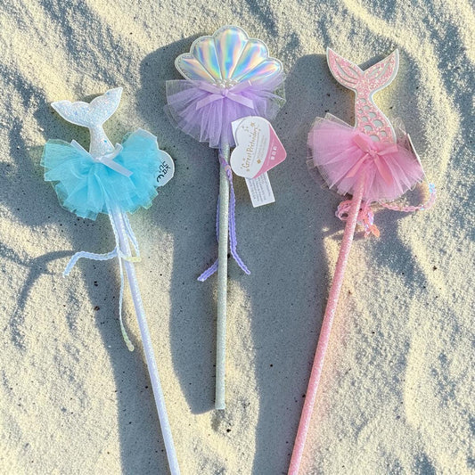 Three mermaid-themed party wands with tutus and tail-shaped tops are arranged on sandy ground in sunlight. The wands are white, pastel purple, and pastel pink, with iridescent and tulle details.