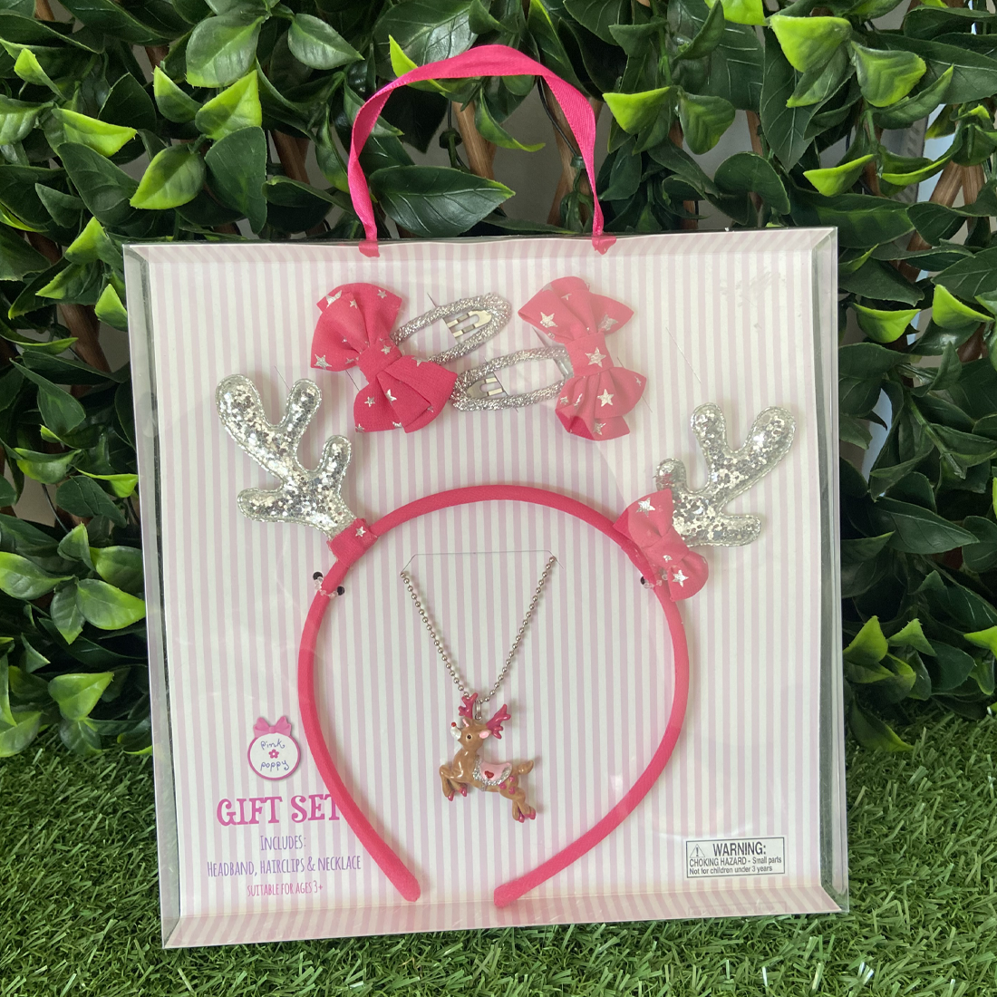 A gift set containing a pink reindeer antler headband, a pink reindeer necklace, two bow hair clips, and a pink headband with silver glitter reindeer antlers and a pink bow featuring metallic stars, packaged in a clear plastic box with a pink ribbon and greenery in the background.
