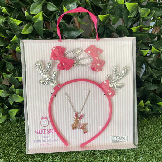 A gift set containing a pink reindeer antler headband, a pink reindeer necklace, two bow hair clips, and a pink headband with silver glitter reindeer antlers and a pink bow featuring metallic stars, packaged in a clear plastic box with a pink ribbon and greenery in the background.