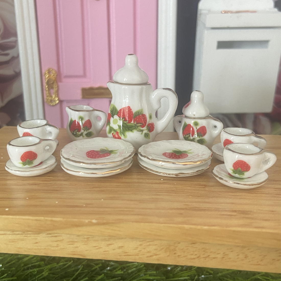 A miniature porcelain tea set with strawberry decorations, including a teapot, milk jug, sugar bowl, plates, and cups, displayed on a wooden surface.