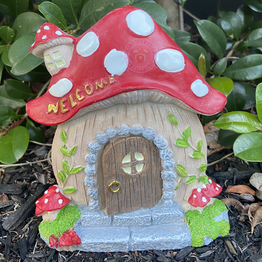 Fairy door with red mushroom-shaped roof and 'Welcome' sign, surrounded by green foliage.