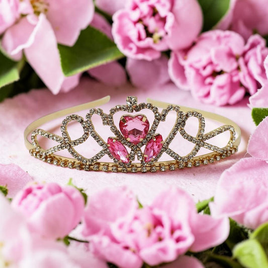 A gold tiara with pink gemstones, styled on a pink surface surrounded by pink flowers.