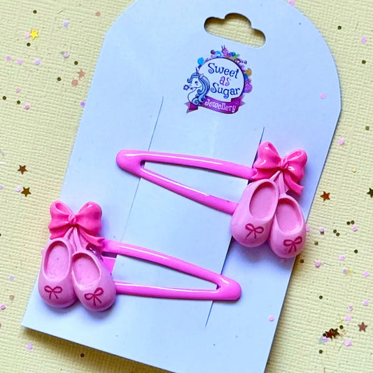 Two pink plastic hair clips shaped like ballet flats with bows, attached to a light blue display card. The card has a 'Sweet As Sugar Jewellery' logo at the top and is photographed on a pale yellow textured surface with scattered gold star confetti.