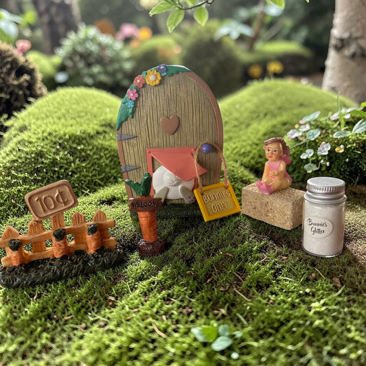Miniature fairy garden scene with a door, sign, and figurines on grass.