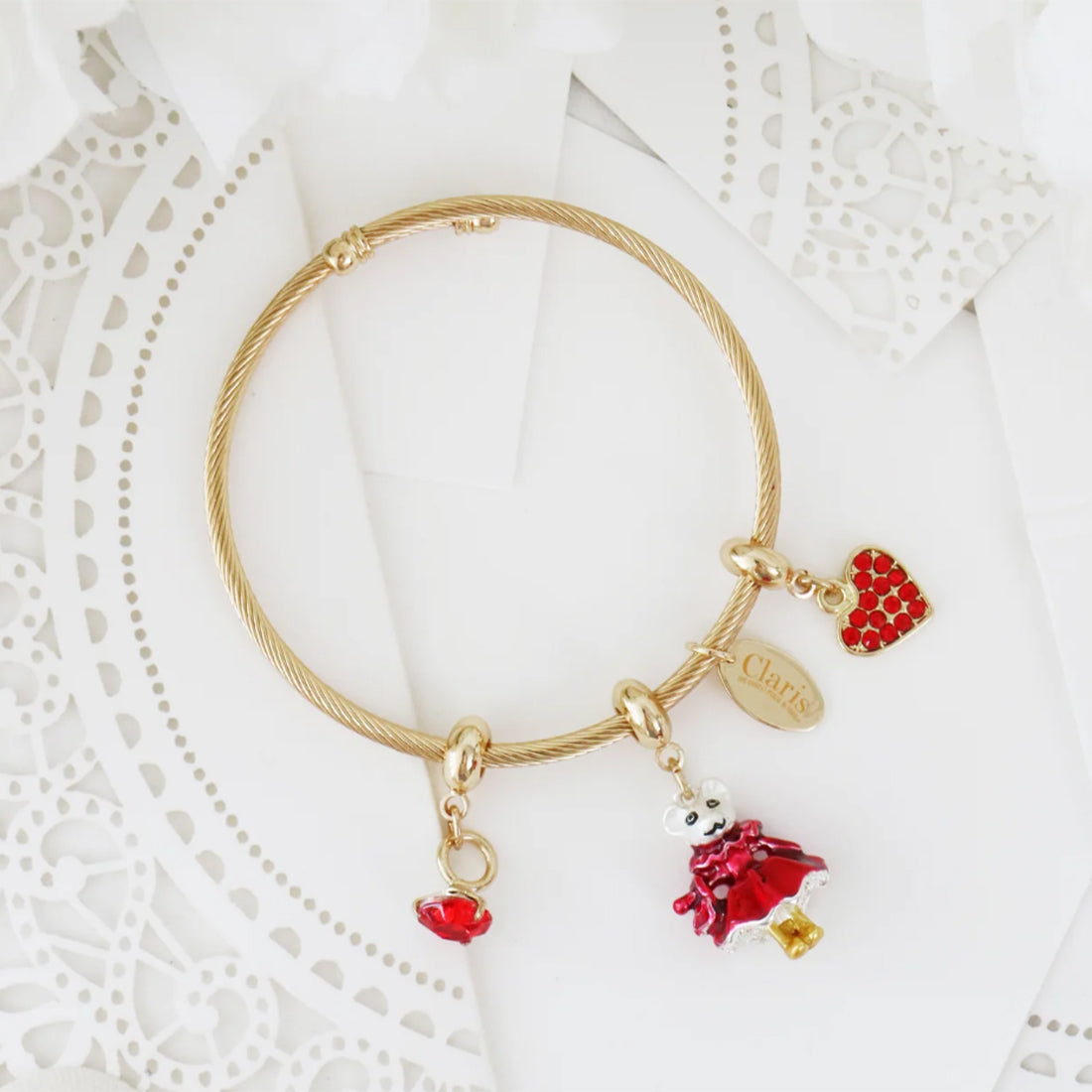 A gold-plated twisted wire bangle bracelet with three dangling claris on paris charms: a red heart charm, a small gold nameplate charm engraved with 'Claris', and a red-and-white strawberry charm, displayed on a white lace doily.