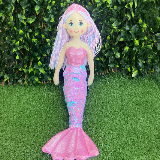 A mermaid doll with long pastel pink and blue braided hair, a pink strapless top, and a shimmering pink mermaid tail, standing on artificial grass in front of a green leafy hedge.