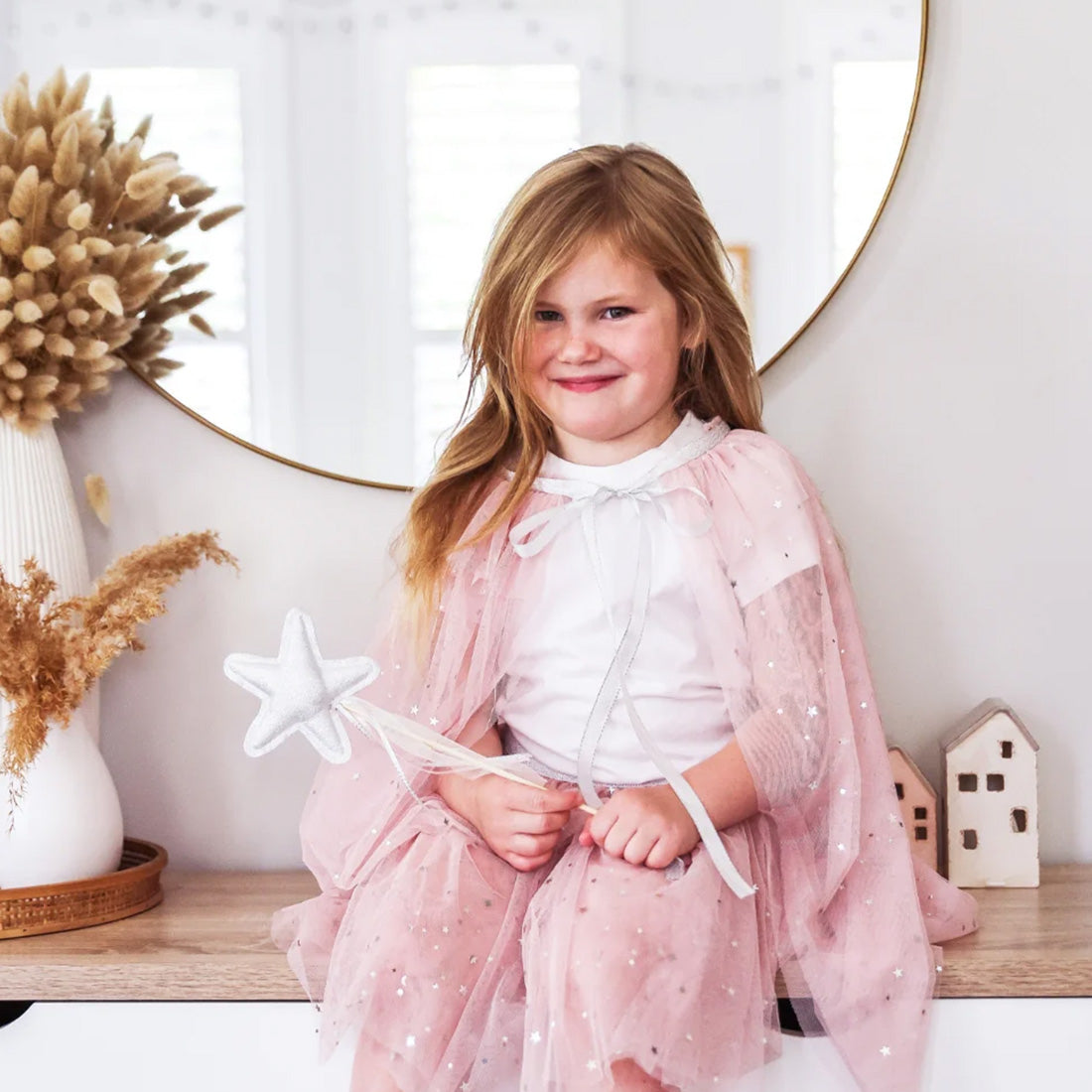 Young girl in a pink cape holding a star-shaped wand in a decorative room.