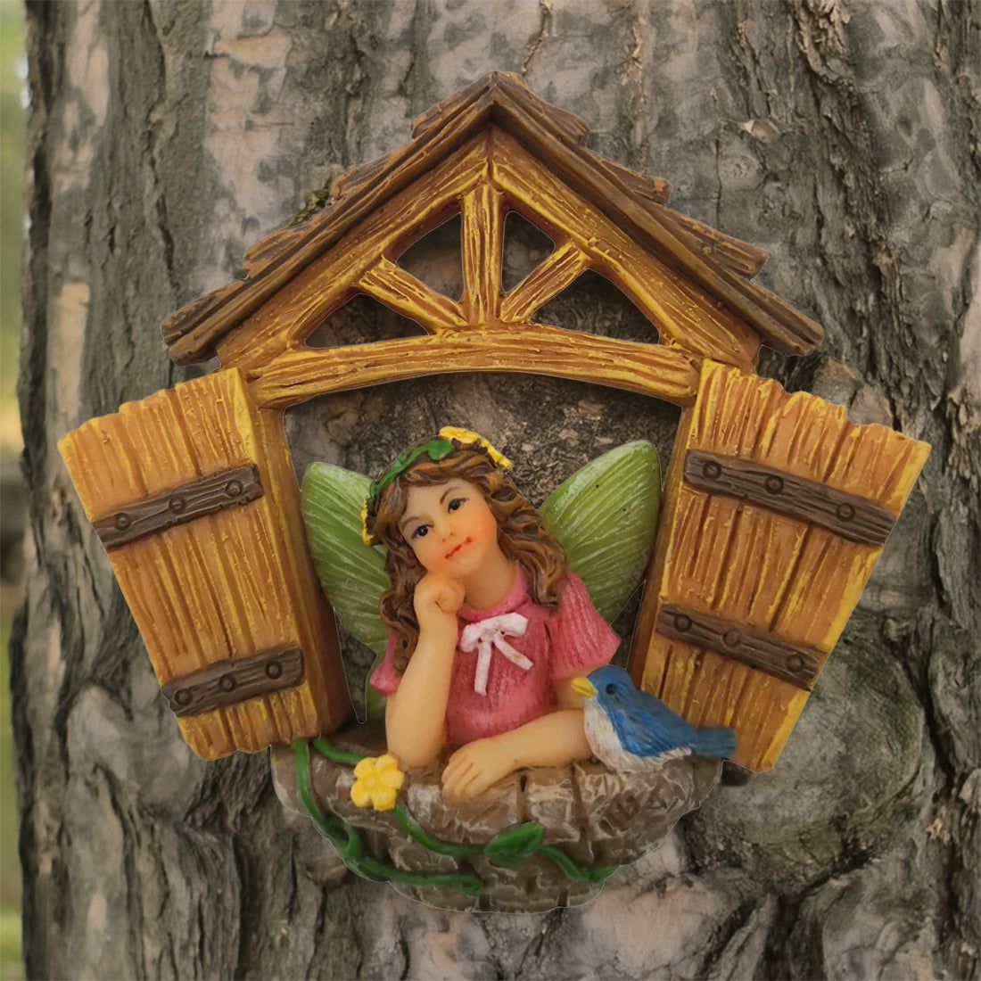 A decorative fairy garden ornament shaped like a small window with open wooden window doors, featuring a day dreaming fairy in a pink dress with green wings, a blue bird, and a yellow flower, mounted on a tree trunk.