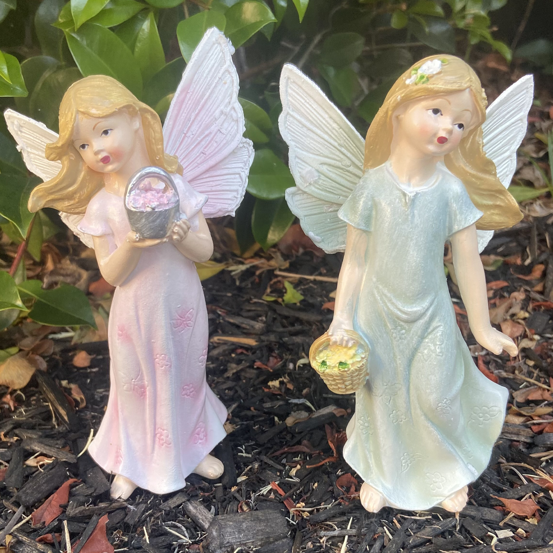 Two angel figurines with wings, one in a pink dress holding a basket, the other in a light blue dress, on a mulched surface.