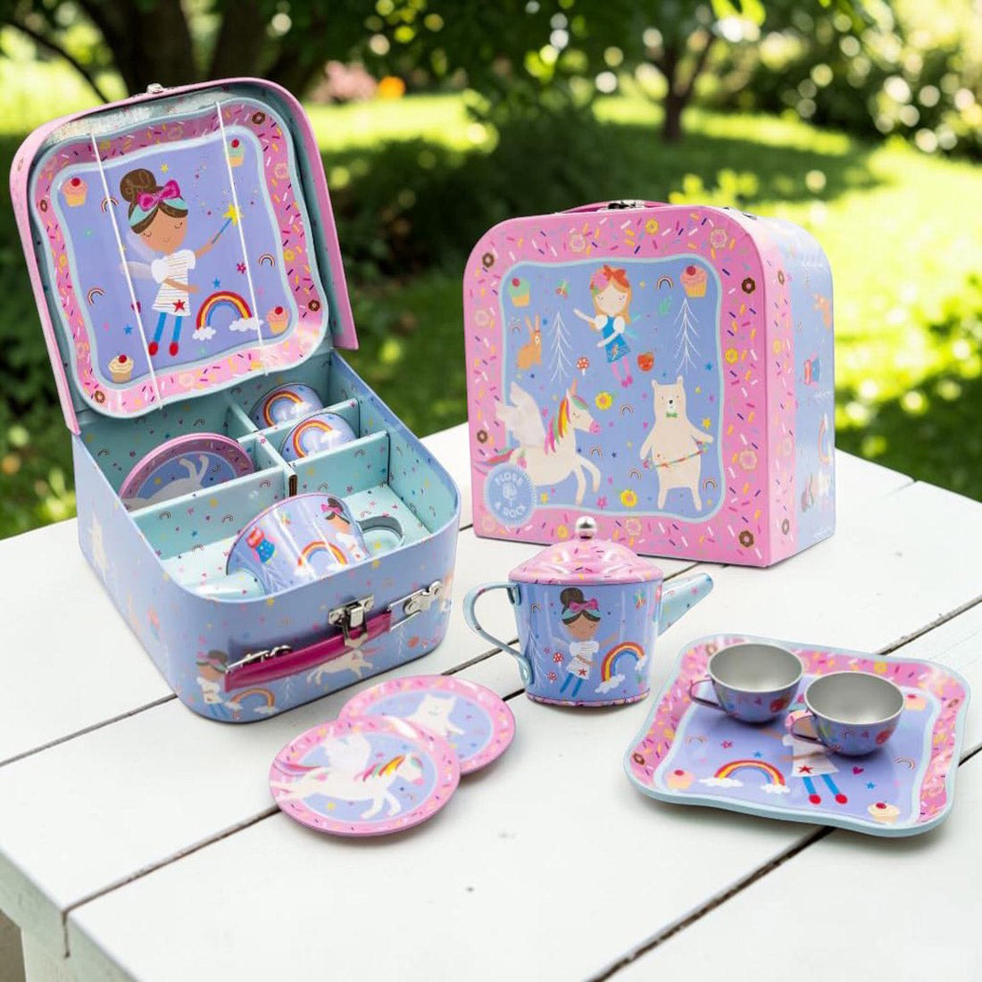A children's tea time set in a pastel blue and pink tin with unicorn and rainbow illustrations, shown open on a white wooden table with matching teapot, cups, saucers, and a box in a garden setting.