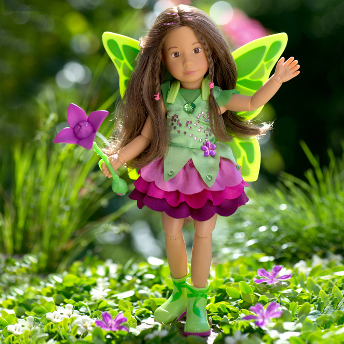 A deluxe fairy doll set featuring a character with brown hair and eyes, wearing a green dress and boots. The set includes a pink dress, a frilly bag, and a hairbrush, posed with a magic flower.