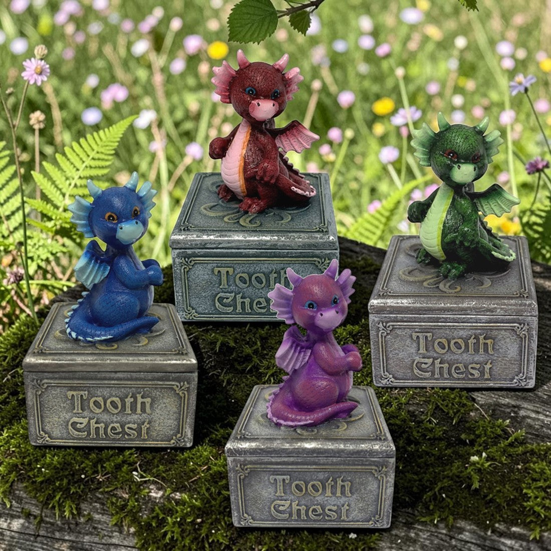 Four small fantasy dragon Tooth Chest figurines displayed outdoors on mossy wood with a blurred wildflower meadow background. The set includes a blue dragon on a gray chest, a dark red dragon on a gray chest, a green dragon on a gray chest, and a purple dragon on a gray chest.