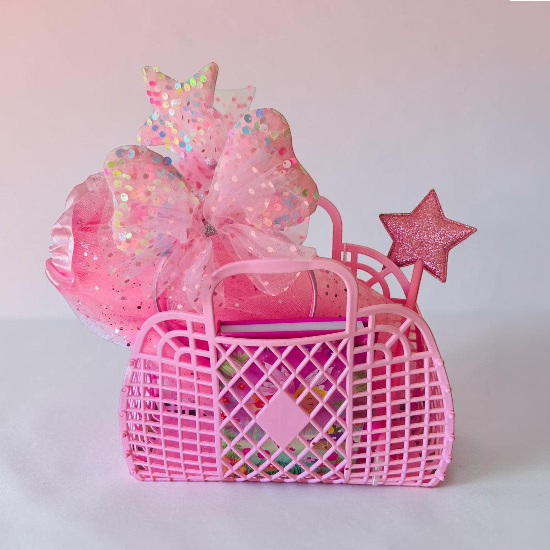 A pink plastic basket gift set filled with colorful items, decorated with a large pink glittery bow, a star topper, and a star wand, set against a soft pink background.