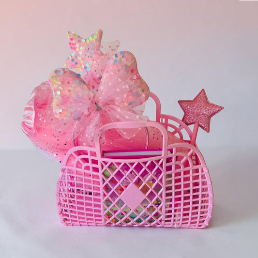 A pink plastic basket gift set filled with colorful items, decorated with a large pink glittery bow, a star topper, and a star wand, set against a soft pink background.