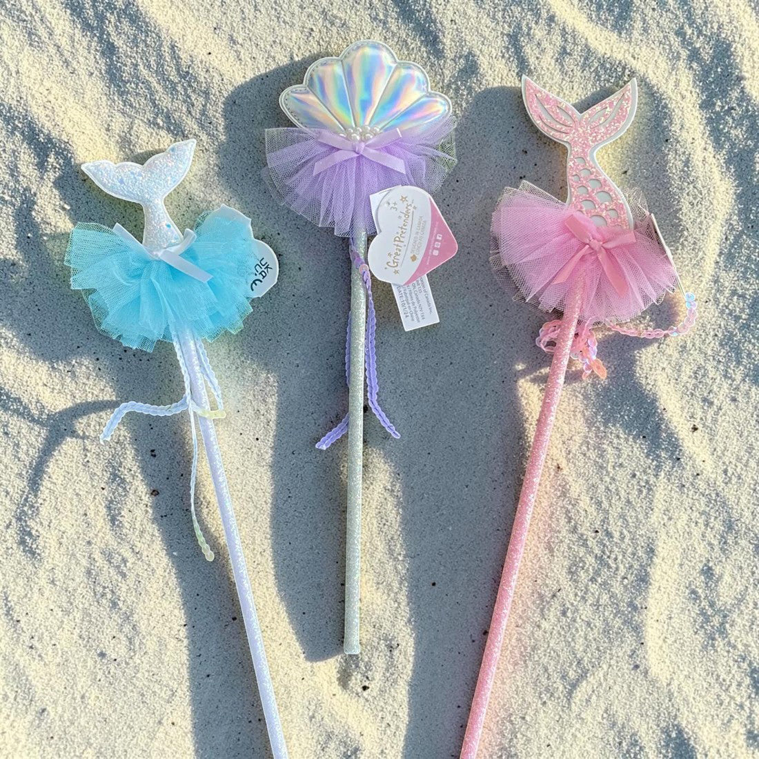 Three mermaid-themed party wands with tutus and tail-shaped tops are arranged on sandy ground in sunlight. The wands are white, pastel purple, and pastel pink, with iridescent and tulle details.