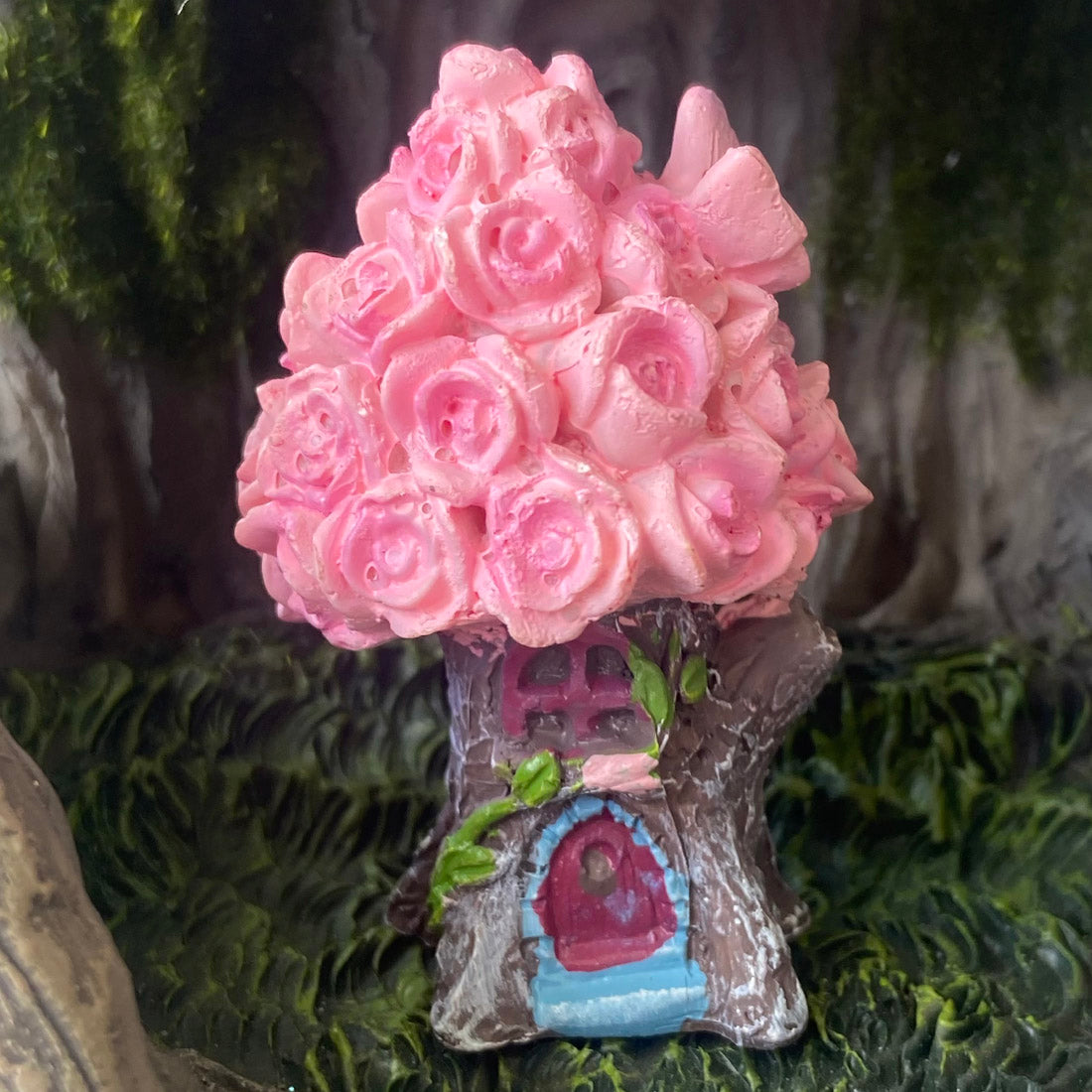 A miniature fairy house shaped like a tree trunk, decorated with a large pink rose-shaped canopy and a small blue and pink door, set against a dark forest background.