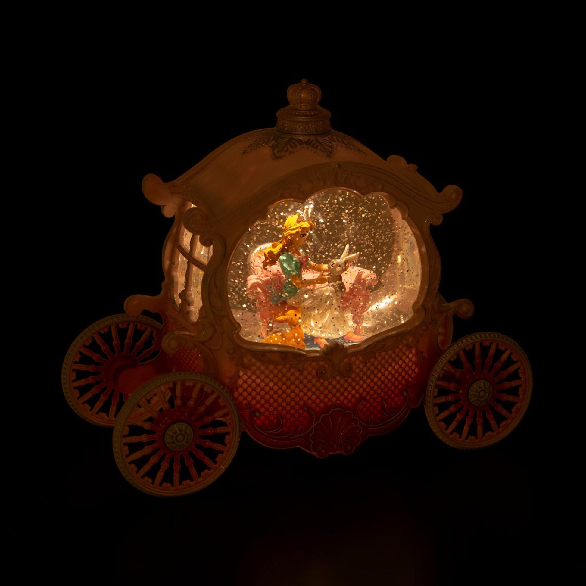 A glowing carriage-shaped snow globe decoration with a warm golden frame, red patterned body, and miniature figures inside, shown against a black background.