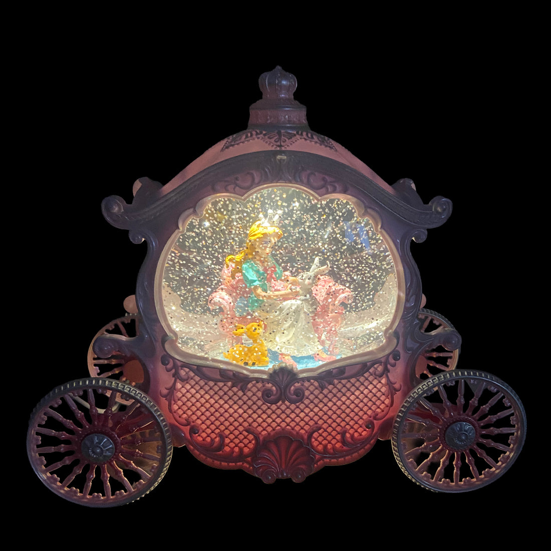 A decorative carriage-shaped music box or snow globe ornament with illuminated gold glitter inside and a small fairy figure, shown against a solid black background.