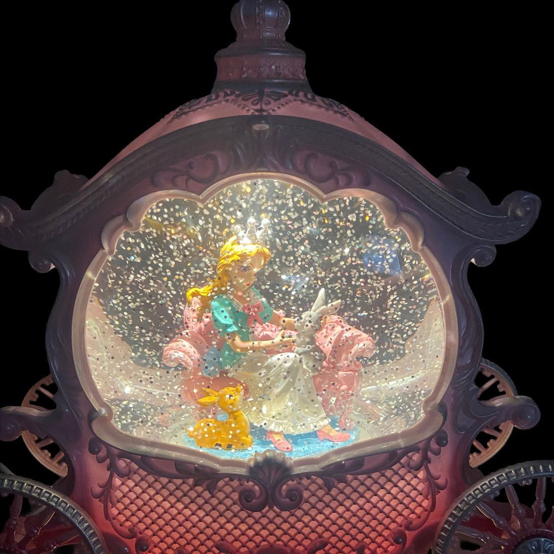 An ornate carriage-shaped snow globe or decorative display case with a glowing interior scene featuring a blonde figure in a pink dress, a white rabbit, and a yellow duck, set against a black background.