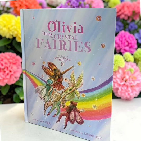 The hardcover children's book 'Olivia Helps the Crystal Fairies' by Jane Prior, featuring a colorful illustration of fairies and a rainbow on a pastel sky cover, displayed on a white surface with blurred pink flowers in the background.