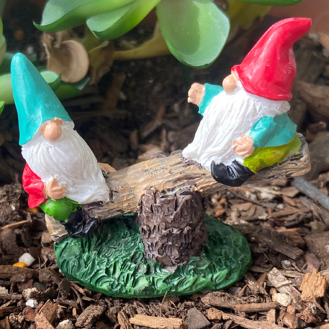 Two miniature gnomes on a log with a red and green hat in a garden setting.